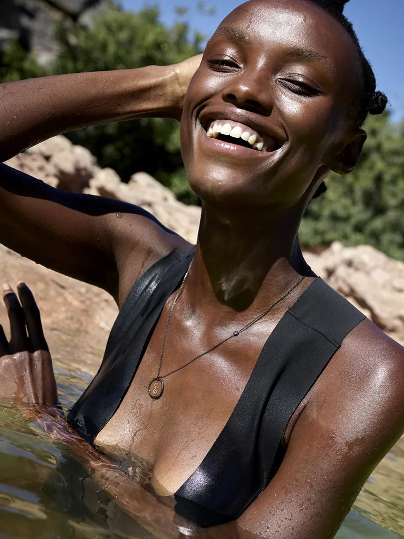 Élégance sous le soleil : Nondi se pavane dans l'eau, sourire rayonnant, avec un pendentif en or noir mettant en valeur l'emblématique symbole de la marque.