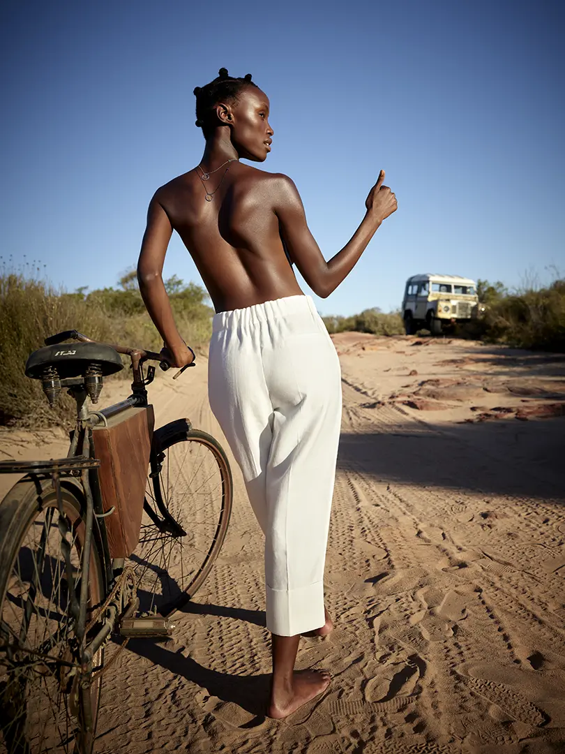 L'aventure de Nondi dans le désert : à vélo, elle cherche son chemin sous le soleil brûlant. Les colliers en Or noir et Or blanc, portés avec élégance dans son dos, ajoutent une touche de glamour à sa quête.