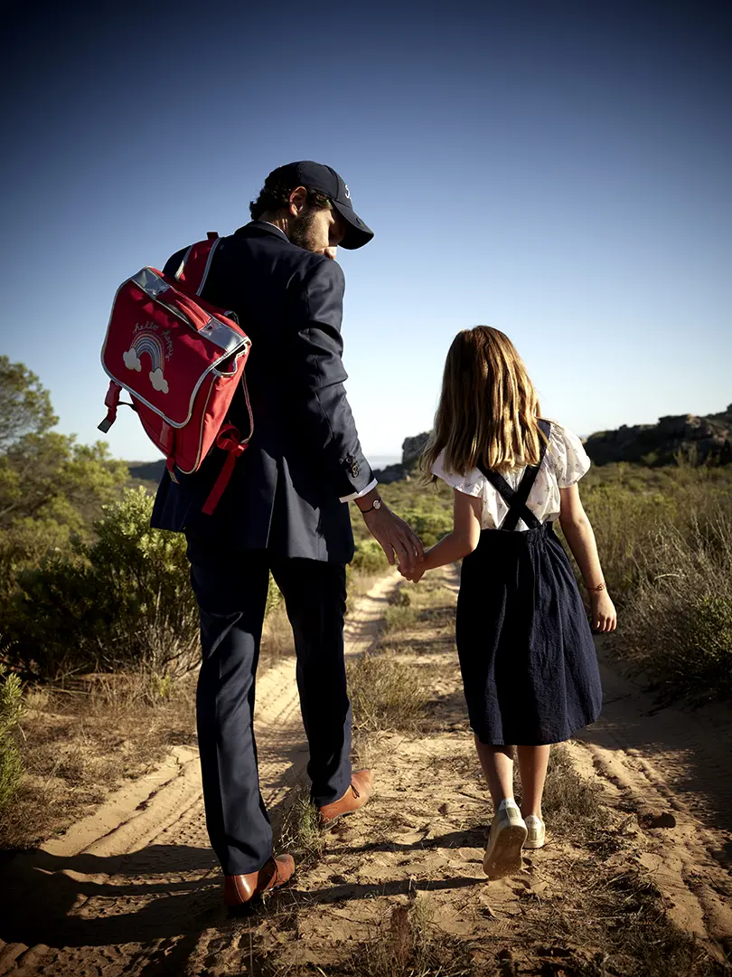 Une traversée scolaire aventureuse : Père et fille, cartable à l'épaule, parcourent les étendues sauvages de la savane avec élégance, portant fièrement un médaillon Cosmos Circle.